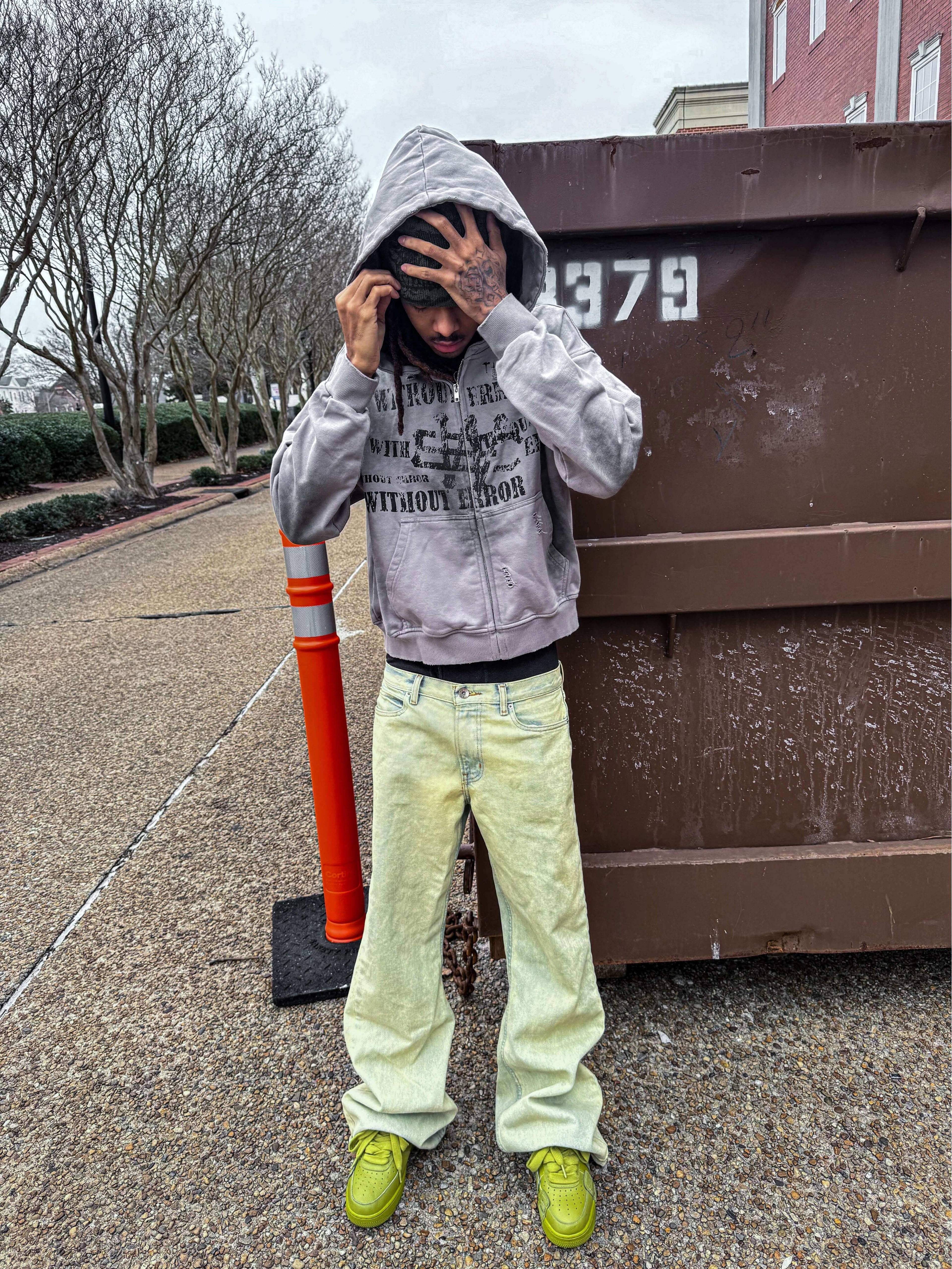 Person wearing a gray hoodie and yellow jeans, standing outdoors near a dumpster, with trees in the background.