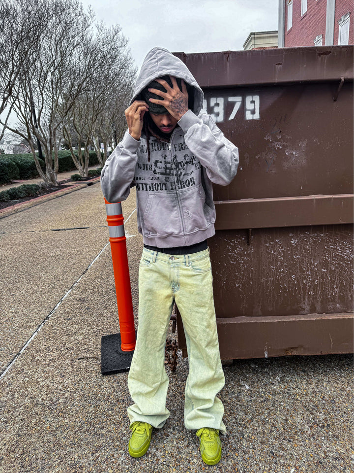 Person wearing a gray hoodie and yellow jeans, standing outdoors near a dumpster, with trees in the background.