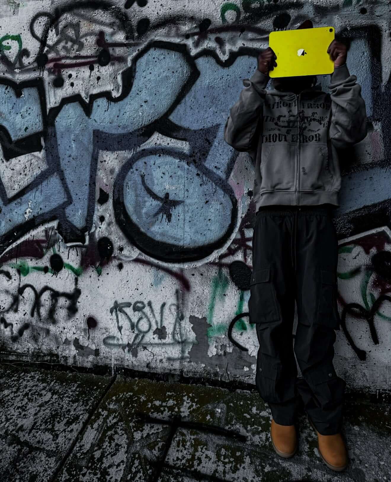 Person wearing a hoodie and cargo pants, holding a yellow tablet in front of a graffiti-covered wall.