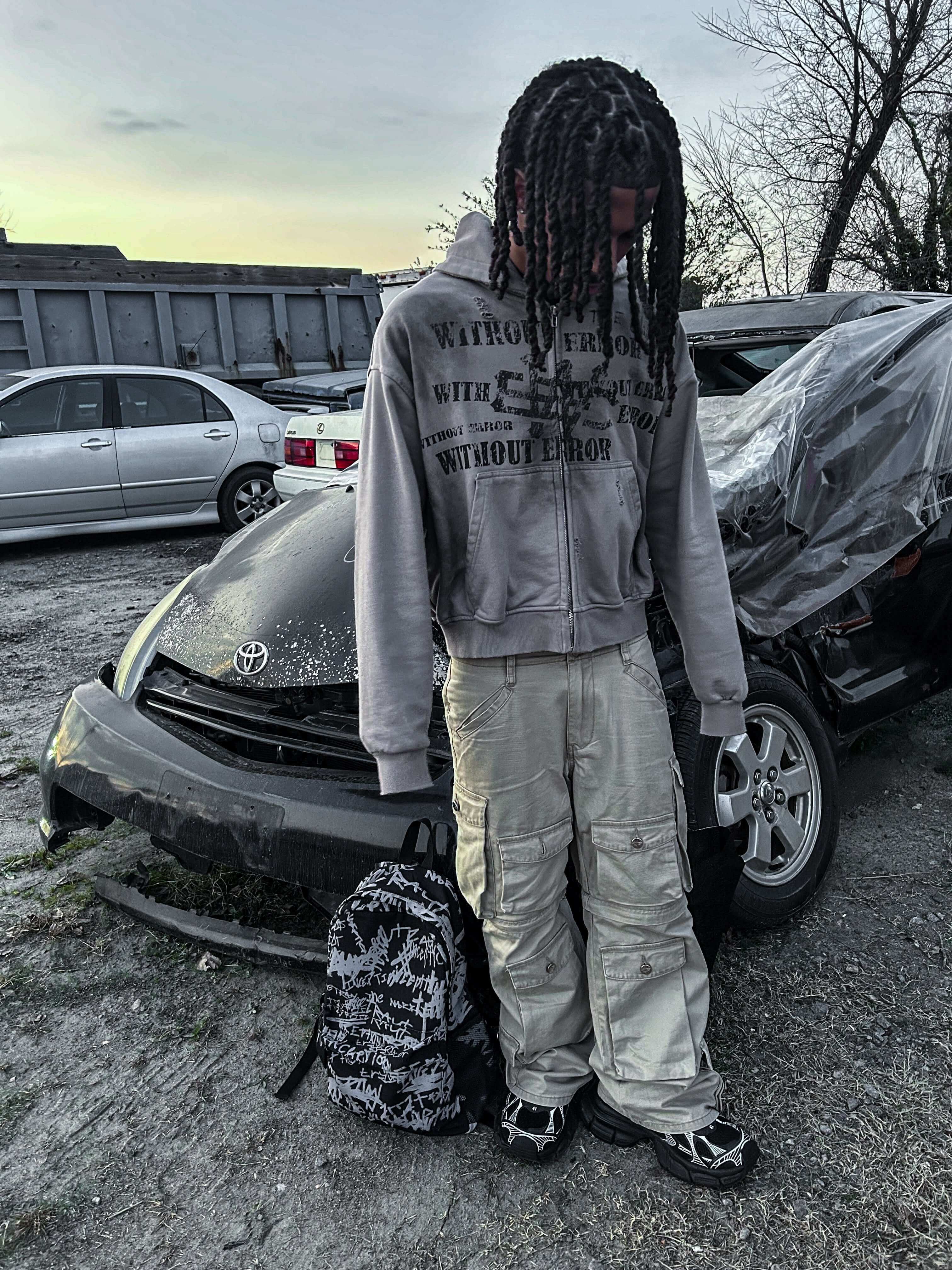 A young person with braided hair stands by a parked car, wearing a hoodie and cargo pants, with a backpack beside them.