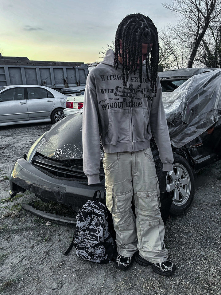 A young person with braided hair stands by a parked car, wearing a hoodie and cargo pants, with a backpack beside them.