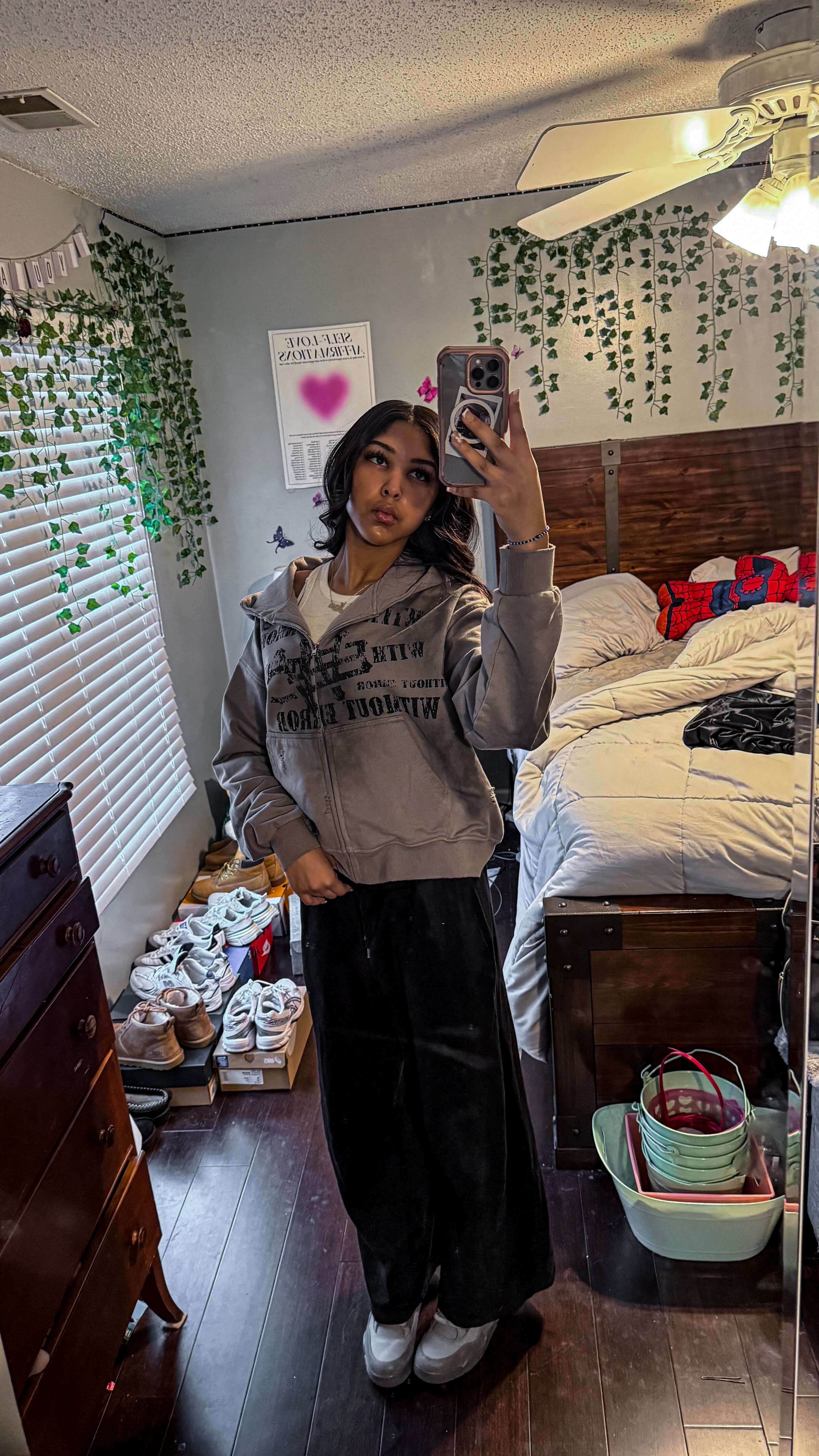 A young woman taking a mirror selfie in a cozy room with plants, wearing a grey hoodie and black pants.