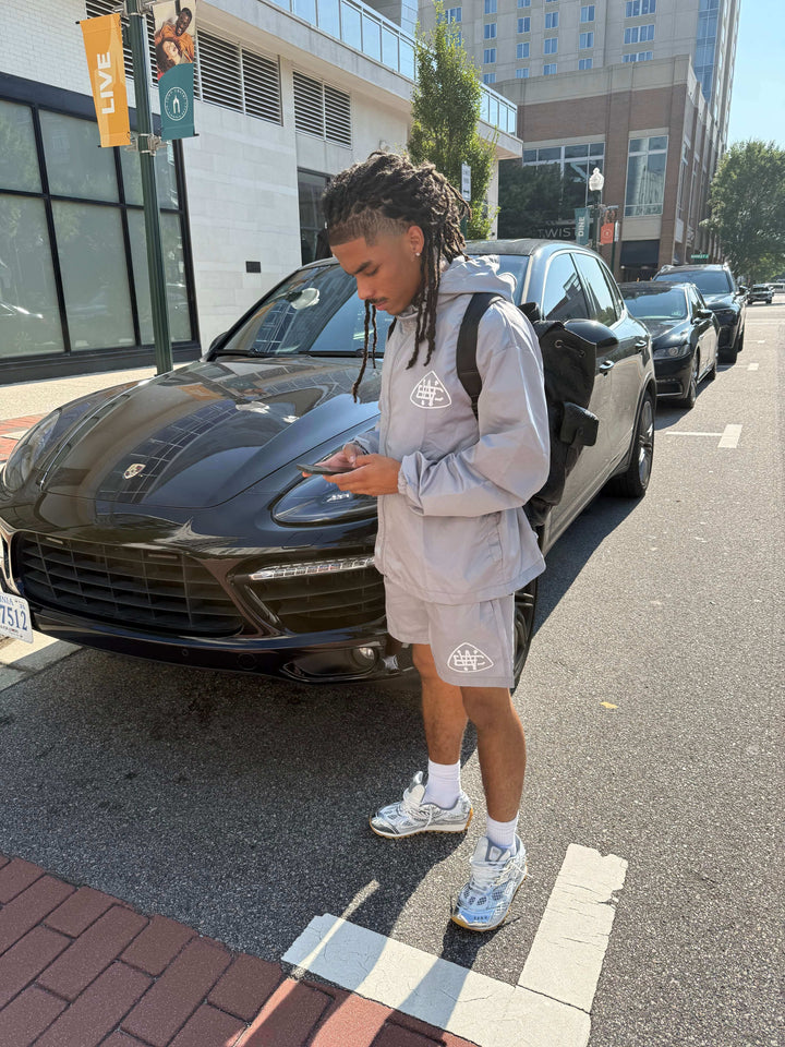 Person wearing a grey drawstring hooded set with a bold logo, standing next to a black Porsche, checking their phone.